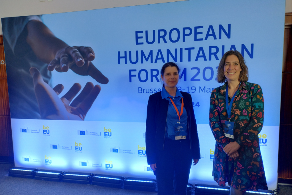 Photo of Gyöngyi Kovács (Hanken) and Claire Barnhoorn (Solvoz) at the European Humanitarian Forum 2024 in front of a forum poster.